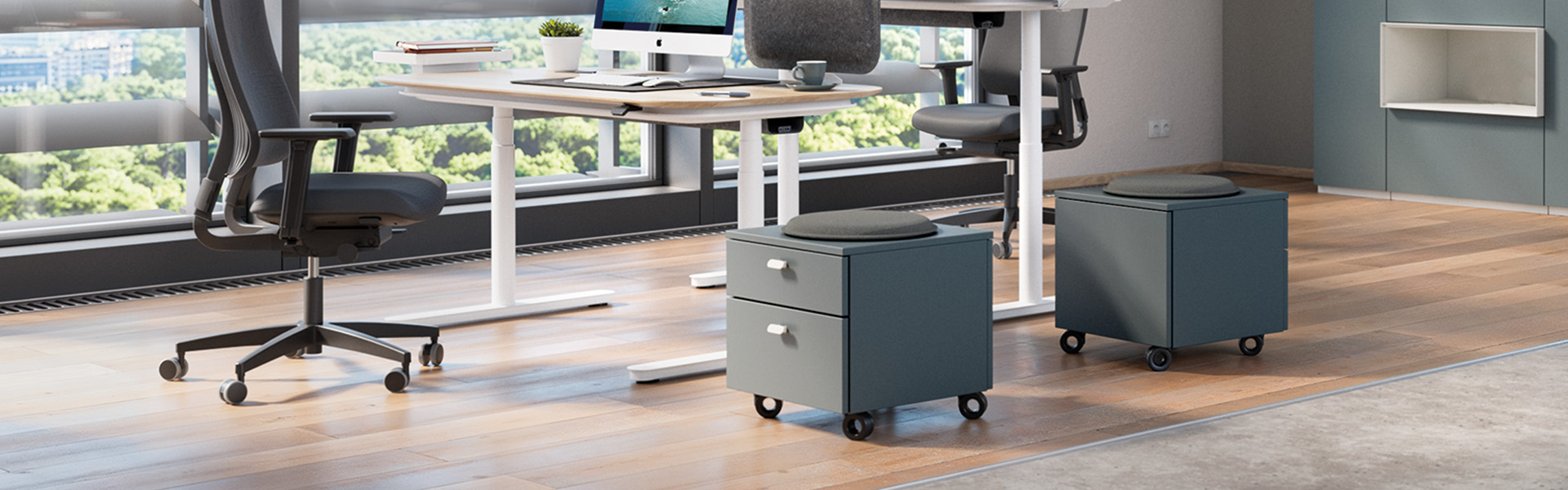 Zwei moderne, dunkelgraue Rollcontainer mit zwei Schubladen und einem runden grauen Sitzkissen darauf, platziert auf einem Büroboden aus Holz neben höhenverstellbaren Schreibtischen und ergonomischen Bürostühlen in einem hellen, modernen Arbeitsberei