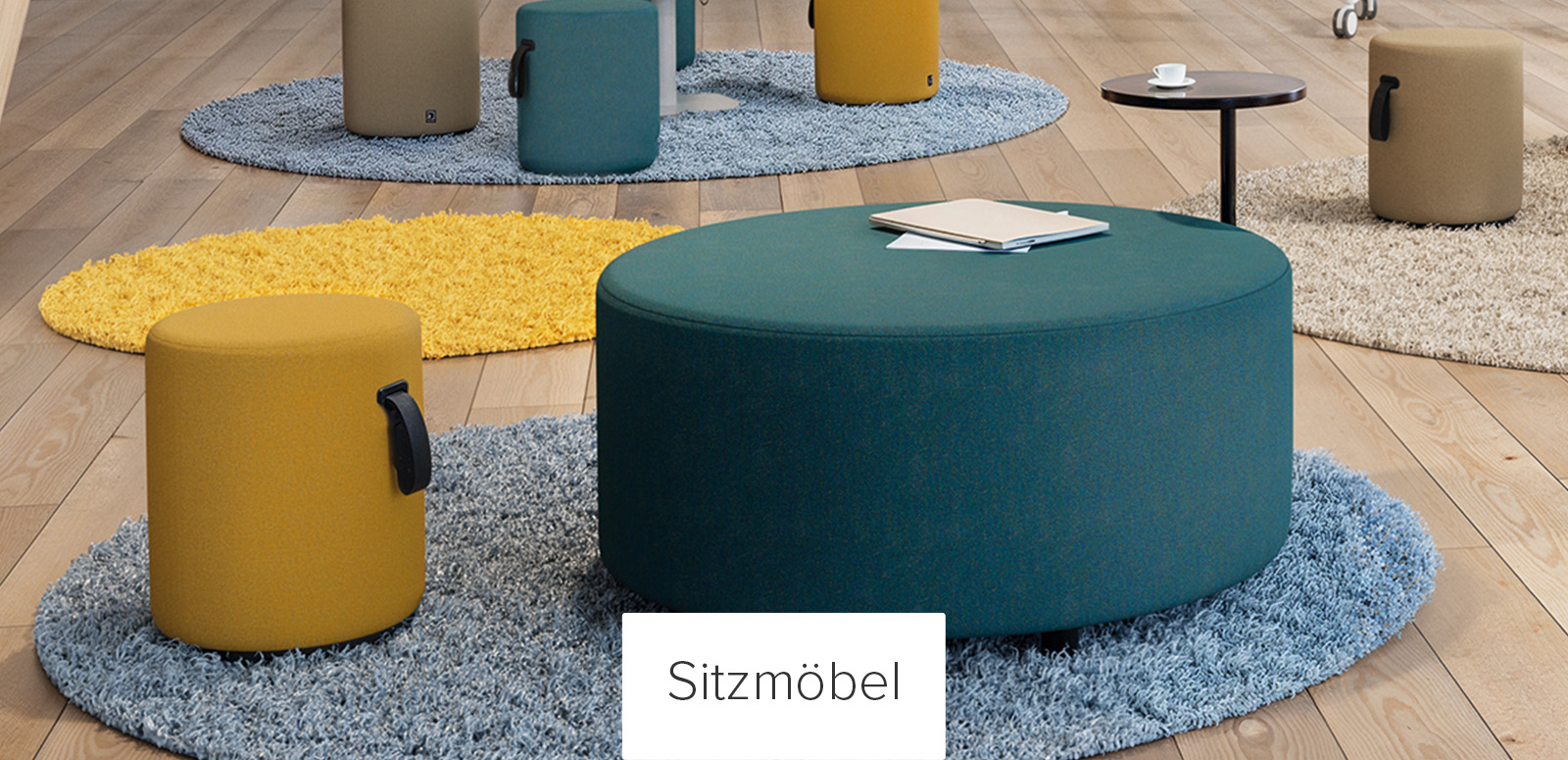 Moderner Büro-Loungebereich mit einer großen runden Ottomane aus türkisfarbenem Stoff auf einem blauen Teppich, umgeben von kleineren zylindrischen Hockern in Senfgelb und Beige mit schwarzen Griffen, auf hellem Holzboden mit farbenfrohen Akzentteppi